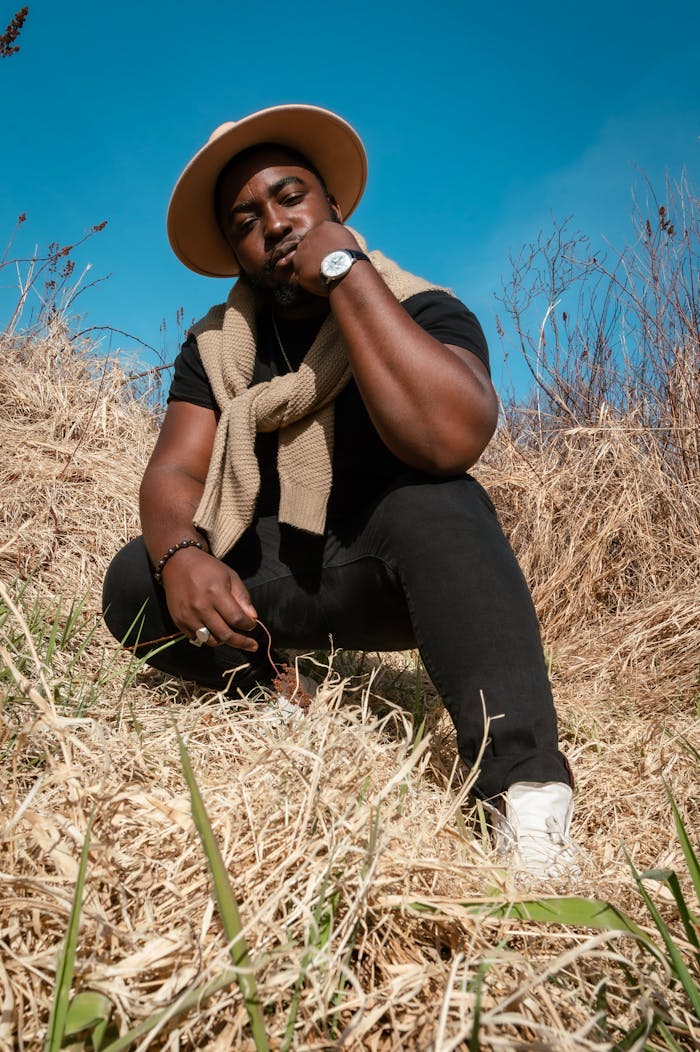 Man in boho style attire sitting in a grassy field under clear blue skies.
