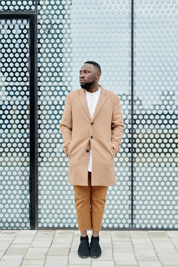 Fashionable man wearing a beige coat stands stylishly against a modern urban backdrop.